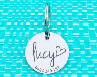 May include: White round pet ID tag with a silver ring. The tag has the name "Lucy" written in cursive with a heart symbol and the phone number "0424 240 183" printed below.