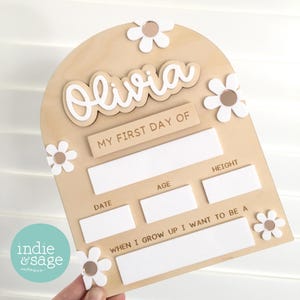 Op de afbeelding: Een houten bord met de naam "Olivia" in witte letters, versierd met witte madeliefjesaccenten. Het bord bevat vakken om details voor "Mijn Eerste Schooldag" vast te leggen, inclusief datum, leeftijd, lengte en ambities.