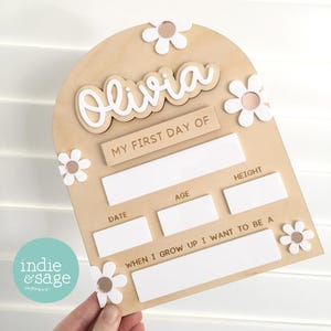 Op de afbeelding: Houten bord voor de eerste schooldag met de naam "Olivia" in witte letters. Het bord heeft een lichtbruine achtergrond, witte madeliefjesaccenten en invulvakken.