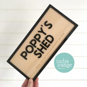 May include: A rectangular wooden sign with a black border and the words "POPPY'S SHED" in bold black letters. Below, smaller text reads "WHERE TOYS ARE FIXED AND MEMORIES ARE MADE." A teal circle with the words "indie & sage" is in the lower right.