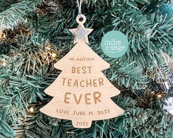 May include: A wooden Christmas tree ornament with the text "Best Teacher Ever" and "Love Jude M. 2023" on it. The ornament is decorated with a silver glitter star.