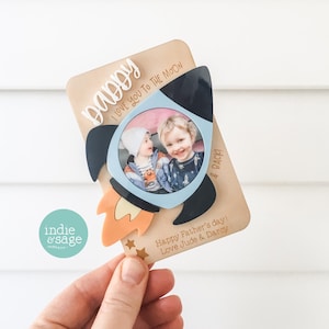 May include: A wooden card with a rocket ship design and a photo of two children. The card says "Daddy I love you to the moon" and "Happy Father's Day! Love Jude & Darcy".