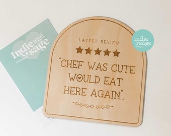 Puede incluir: Un letrero de madera con la parte superior redondeada, que presenta una calificación de cinco estrellas y el texto "LATEST REVIEW" sobre la cita "CHEF WAS CUTE WOULD EAT HERE AGAIN." El letrero está decorado con pequeños corazones.