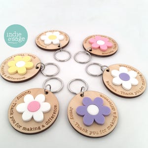 May include: Six circular wooden keychains with metal rings, each featuring a raised flower shape in various colours including white, pink, yellow, and purple. The wood is engraved with the text Thank you for making a difference and a name.