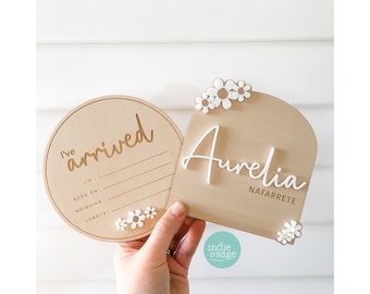 Puede incluir: Dos letreros de madera para anunciar el nacimiento de un bebé. Uno es redondo con las palabras "I've arrived" y espacio para detalles. El otro es arqueado con el nombre "Aurelia" y "Nafarrete". Ambos tienen detalles de margaritas.