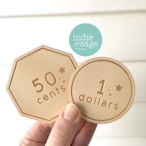 May include: Two wooden play money tokens are held in a hand. One is a light brown octagon with "50 cents" engraved. The other is a round token with "1 dollars" engraved. Both have star accents.