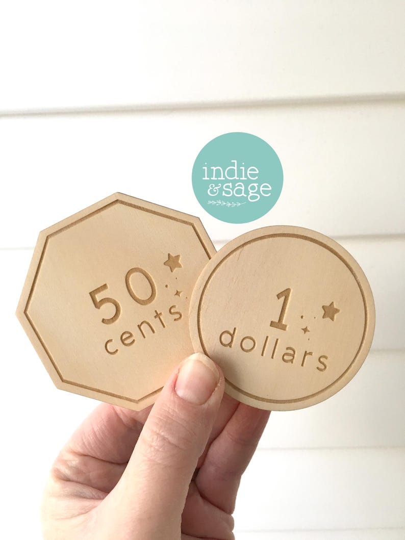 May include: Two wooden play money tokens are held in a hand. One is a light brown octagon with "50 cents" engraved. The other is a round token with "1 dollars" engraved. Both have star accents.