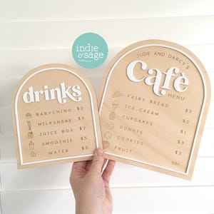 May include: Two wooden cafe menu signs with white lettering and borders. One sign says "drinks" with a list of beverages and prices. The other says "Jude and Darcy's Cafe Menu" with food items and prices.