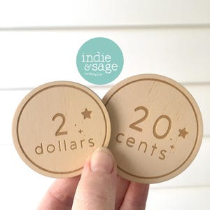 May include: Two wooden play money coins. One coin reads "2 dollars" and the other reads "20 cents". Both coins have a star and small starburst design. The brand logo "indie & sage" is visible in the background.
