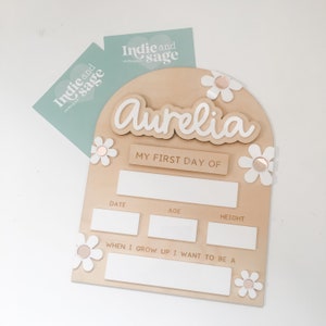 Op de afbeelding: Een houten bord met de naam "Aurelia" in witte letters, versierd met witte madeliefjesaccenten. Het bord bevat vakken voor "Datum", "Leeftijd" en "Lengte", en de tekst "My First Day Of". Twee mintgroene kaarten met het logo "Indie and sage" op de achtergrond.