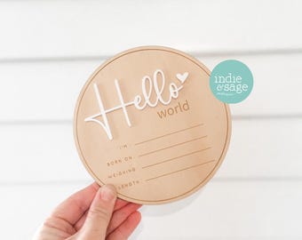 Puede incluir: Un letrero redondo de madera con el texto "Hello World" en escritura blanca con un pequeño corazón. El letrero tiene líneas en blanco para "Soy", "Nacido el", "Peso", y "Longitud".