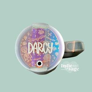 May include: A white personalized tumbler with a clear lid featuring a Halloween-themed design with bats, ghosts, and skeletons. The name "Darcy" is written in white on the lid.