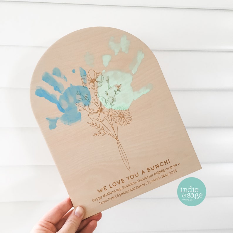 May include: Wooden plaque with an arched top, featuring a hand-painted blue handprint and a bouquet of flowers. The text "WE LOVE YOU A BUNCH!" is engraved, along with a Mother's Day message and names. The plaque is held by a hand.