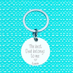 Puede incluir: Llavero de plata con una etiqueta redonda que dice "El mejor pap&aacute; me pertenece" con un s&iacute;mbolo de coraz&oacute;n y el nombre "Liam" debajo.