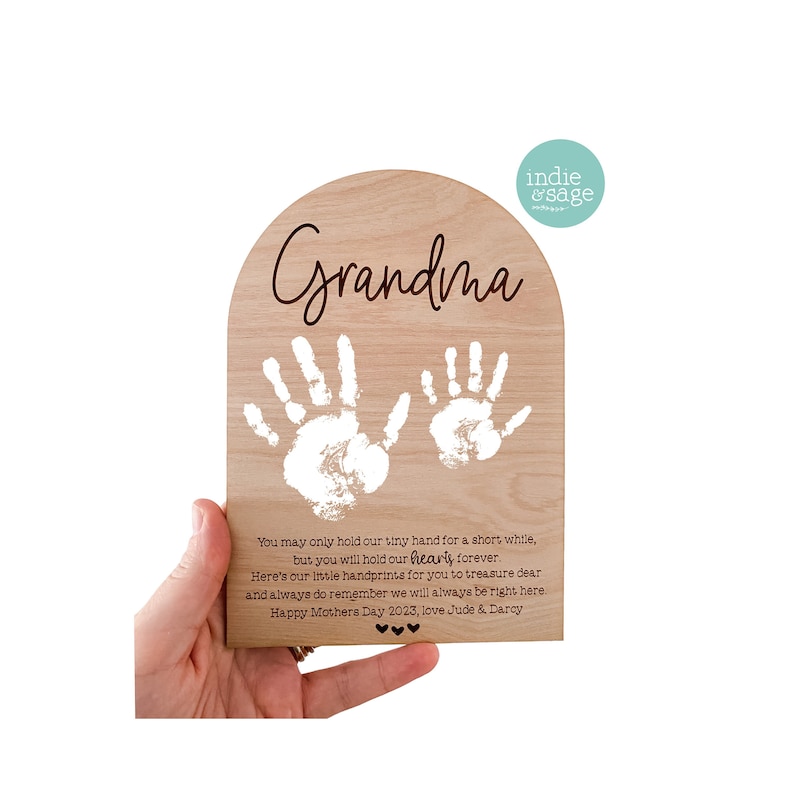 K&ouml;nnte beinhalten: Holztafel mit hellem Holzmaserung, mit dem Wort "Oma" in Schreibschrift. Zwei wei&szlig;e Handabdr&uuml;cke befinden sich darunter. Die Tafel enth&auml;lt eine herzliche Botschaft und den Text "Happy Mothers Day 2023, love Jude & Darcy."