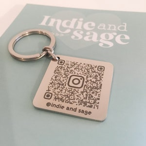 May include: A silver keychain with a square metal tag featuring a black and white QR code with an Instagram logo in the centre. The text "@indie and sage" is engraved below the QR code.