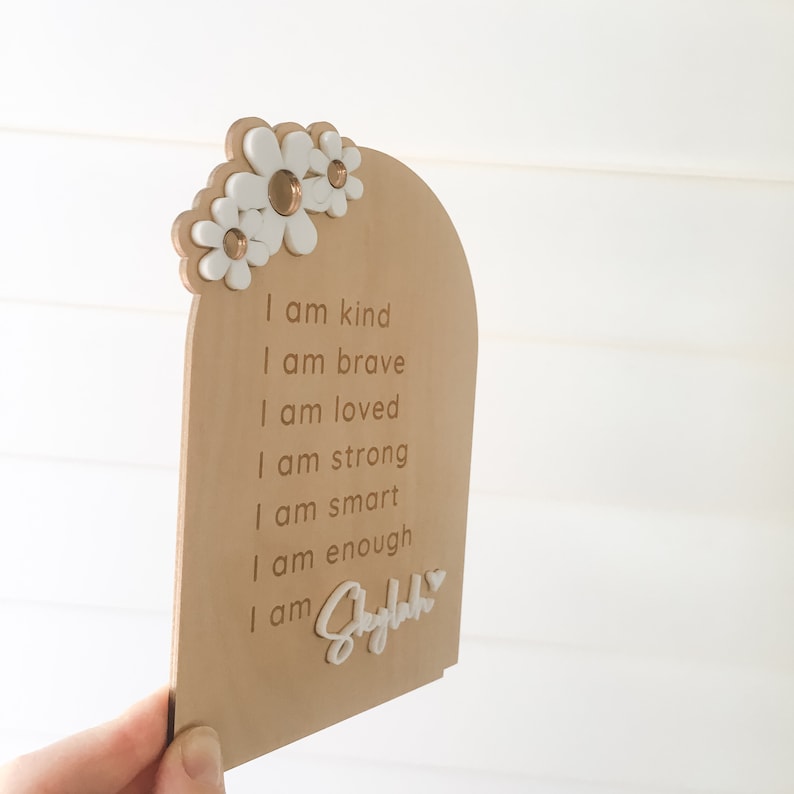 Puede incluir: Un letrero de madera con forma de arco blanco y flores blancas en la parte superior. El letrero tiene el texto "Soy amable, soy valiente, soy amado, soy fuerte, soy inteligente, soy suficiente, soy Skylah" escrito en &eacute;l.