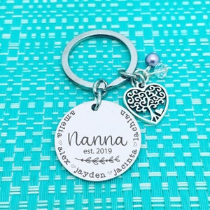 May include: Silver keychain with a round tag that says "Nanna est. 2019" with the names Amelia, Alex, Jayden, and Jacinta around the edge. A silver heart charm with a tree design is attached to the keychain.