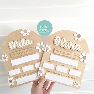Op de afbeelding: Twee houten borden voor de eerste schooldag met de namen "Mila" en "Olivia". Elk bord heeft een madeliefjesontwerp en vakken voor datum, leeftijd en lengte. De borden zijn lichtbruin.