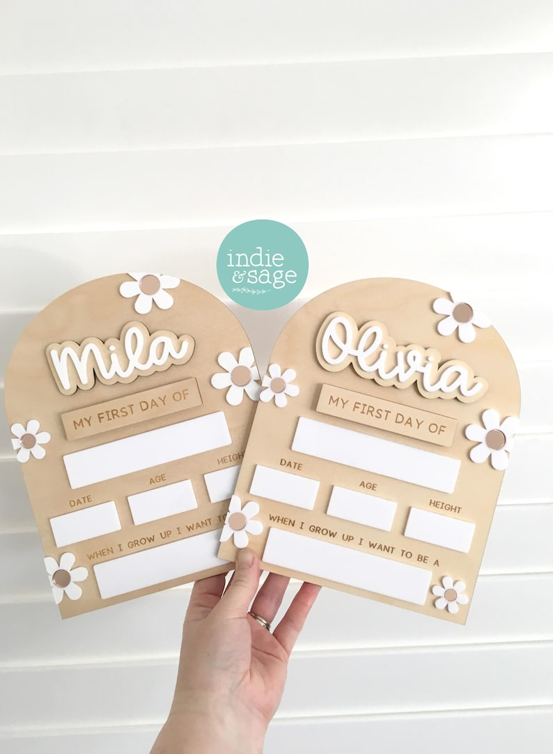 Op de afbeelding: Twee houten borden voor de eerste schooldag met de namen "Mila" en "Olivia". Elk bord heeft een madeliefjesontwerp en vakken voor datum, leeftijd en lengte. De borden zijn lichtbruin.