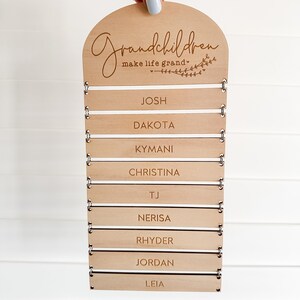 May include: A wooden sign with the words "Grandchildren make life grand" engraved at the top. Below are slots for name tags, with names like Josh, Dakota, and Christina. The sign is held by a hand with a light blue nail polish.