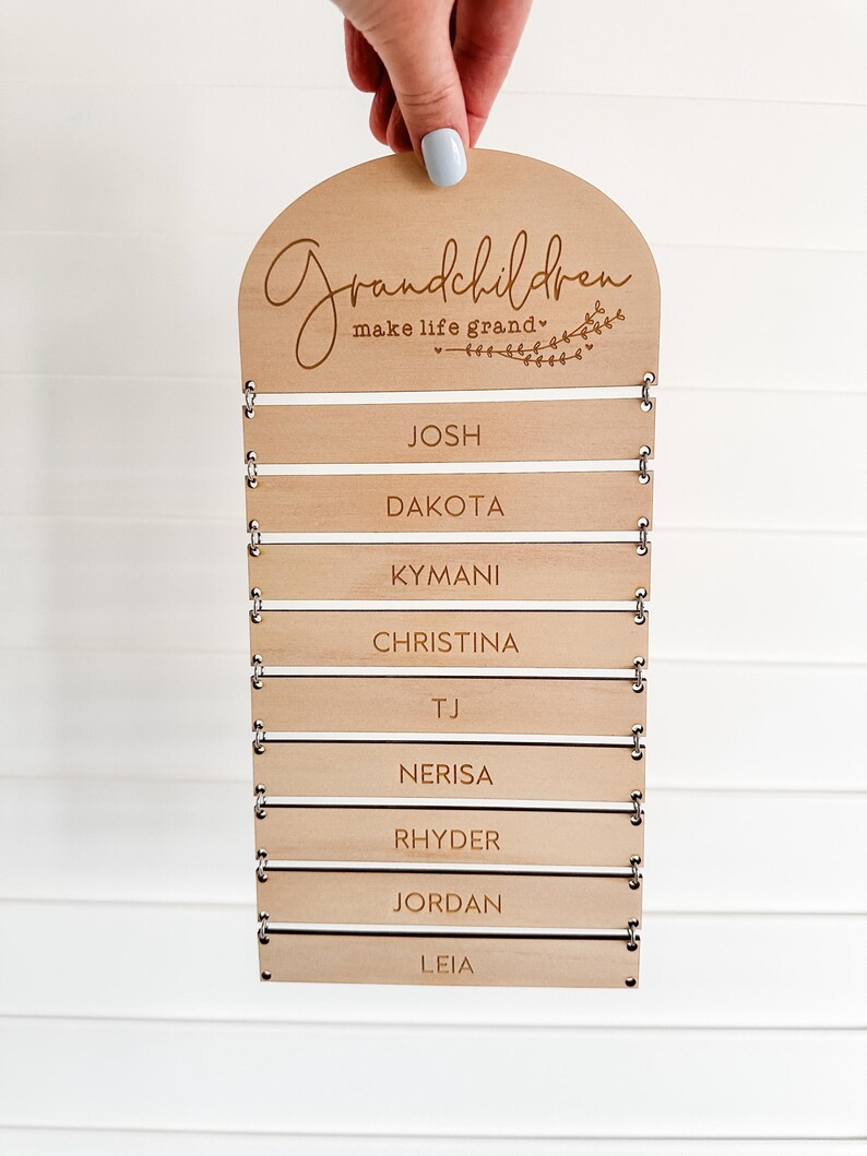 May include: A wooden sign with the words "Grandchildren make life grand" engraved at the top. Below are slots for name tags, with names like Josh, Dakota, and Christina. The sign is held by a hand with a light blue nail polish.