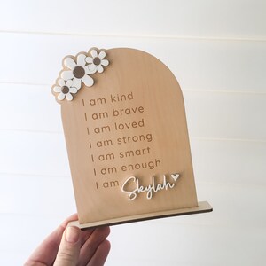 Puede incluir: Un letrero de madera con la parte superior redondeada y un soporte. El letrero tiene un dise&ntilde;o de margarita blanca y el texto "Soy amable, soy valiente, soy amada, soy fuerte, soy inteligente, soy suficiente, soy Skylah" con un s&iacute;mbolo de coraz&oacute;n.