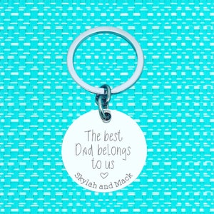 Puede incluir: Un llavero plateado con una etiqueta redonda blanca que dice "The best Dad belongs to us Skylar and Mack" con un s&iacute;mbolo de coraz&oacute;n entre los nombres.