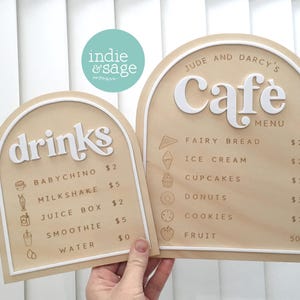 May include: Two wooden cafe menu signs with arched tops. One sign says "drinks" with a list of beverages and prices. The other says "Cafe Menu" with food items and prices. The signs have a light wood grain background with white lettering.