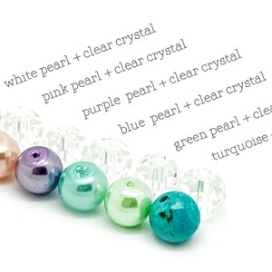 May include: A close-up of a rainbow of pearl beads with clear crystal beads interspersed. The beads are arranged in a line from left to right: white, pink, purple, blue, green, and turquoise.