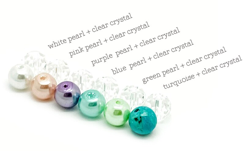 May include: A close-up of a rainbow of pearl beads with clear crystal beads interspersed. The beads are arranged in a line from left to right: white, pink, purple, blue, green, and turquoise.