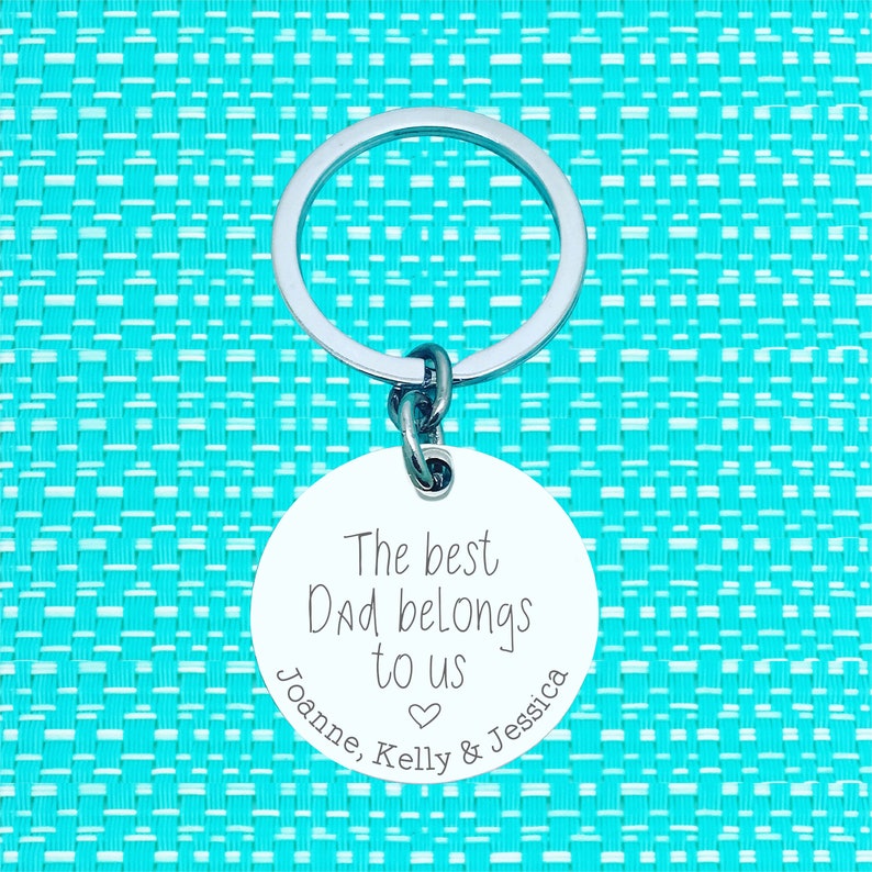 Puede incluir: Un llavero plateado con una etiqueta redonda blanca que dice "The best Dad belongs to us Joanne, Kelly & Jessica" con un s&iacute;mbolo de coraz&oacute;n.