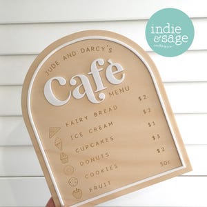 May include: A wooden cafe menu with a light brown surface and a white border. The menu lists items like fairy bread, ice cream, and donuts. The cafe's name, "Jude and Darcy's Cafe," is displayed at the top.