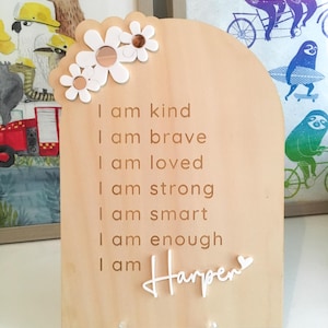 Puede incluir: Un letrero de madera con forma de arco blanco y un dise&ntilde;o floral en la parte superior. El letrero tiene el texto "I am kind, I am brave, I am loved, I am strong, I am smart, I am enough, I am Harper" en letras blancas.