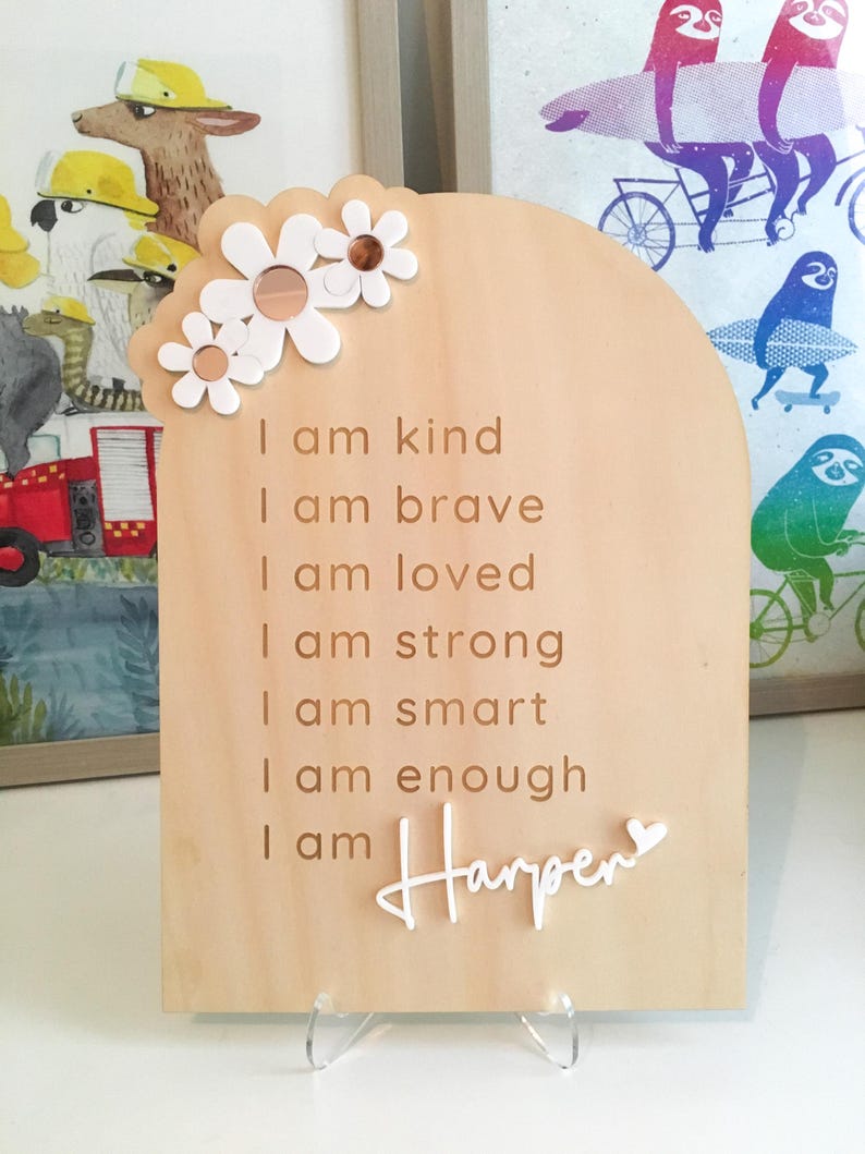 Puede incluir: Un letrero de madera con forma de arco blanco y un dise&ntilde;o floral en la parte superior. El letrero tiene el texto "I am kind, I am brave, I am loved, I am strong, I am smart, I am enough, I am Harper" en letras blancas.