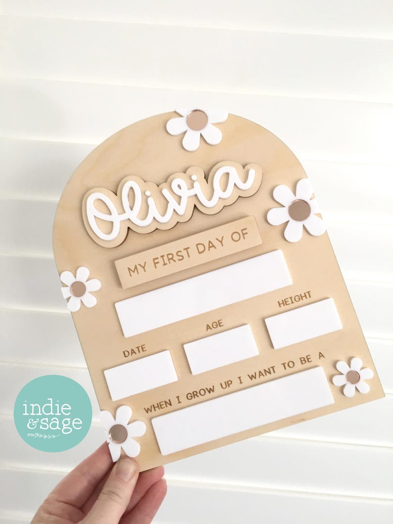 Op de afbeelding: Een houten bord met de naam "Olivia" in witte letters, versierd met witte madeliefjesaccenten. Het bord bevat vakken om details voor "Mijn Eerste Schooldag" vast te leggen, inclusief datum, leeftijd, lengte en ambities.