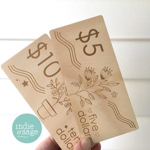 May include: Two wooden gift cards with engraved designs. One card reads "$10" and the other reads "$5". Both cards feature floral and wavy line designs. The "indie & sage" logo is visible in the lower left corner.