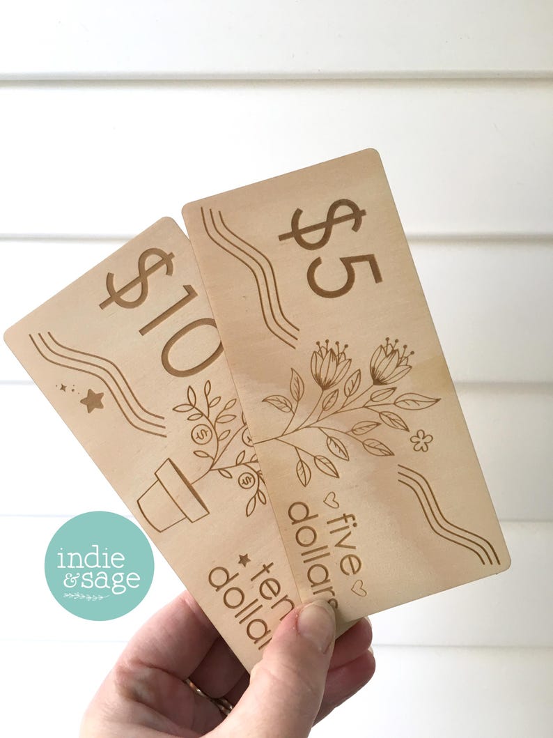 May include: Two wooden gift cards with engraved designs. One card reads "$10" and the other reads "$5". Both cards feature floral and wavy line designs. The "indie & sage" logo is visible in the lower left corner.