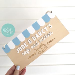 May include: A rectangular wooden sign featuring a light blue and white scalloped awning at the top. The sign displays the white raised text JUDE & DARCY'S cafe and kitchen, accompanied by an engraved illustration of a teacup and leafy accents.
