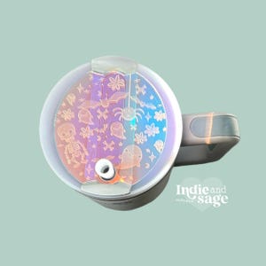 May include: A white travel mug with a clear lid featuring a holographic design of Halloween-themed icons, including bats, ghosts, spiders, and a skeleton. The lid has a small hole for a straw.