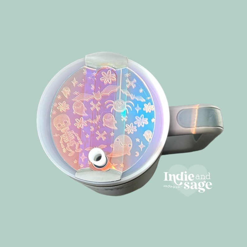 May include: A white travel mug with a clear lid featuring a holographic design of Halloween-themed icons, including bats, ghosts, spiders, and a skeleton. The lid has a small hole for a straw.
