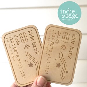 May include: Two wooden kids bank cards with rounded corners. Each card has a name, account number, and the words "Kids Bank" and "Savings Account" engraved. One card is labeled "Jude", the other "Darcy". A star and wave design is also present.