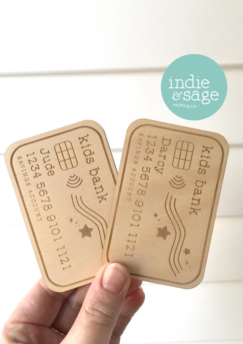 May include: Two wooden kids bank cards with rounded corners. Each card has a name, account number, and the words "Kids Bank" and "Savings Account" engraved. One card is labeled "Jude", the other "Darcy". A star and wave design is also present.
