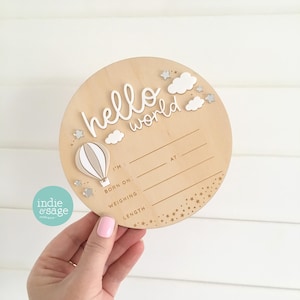 May include: A circular wooden sign with the words "hello world" in white, a hot air balloon, and clouds. The sign has lines for baby's details. The sign is light brown with silver stars and clouds.