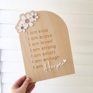 Puede incluir: Un letrero de madera con un fondo marr&oacute;n claro y flores de margarita blancas. El letrero tiene el texto "Soy amable, soy valiente, soy amado, soy fuerte, soy inteligente, soy suficiente, soy Harper" en letras doradas.