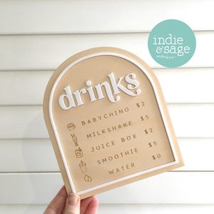 May include: A tan-colored drinks menu with a white border and the word "drinks" in a stylized font. The menu lists drink options including Babychino, Milkshake, Juice Box, Smoothie, and Water.