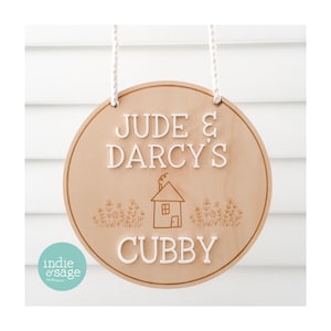 May include: A round wooden sign with the text "Jude & Darcy's Cubby" in white letters. The sign has a small house and flowers engraved on it and is hanging from a white rope.