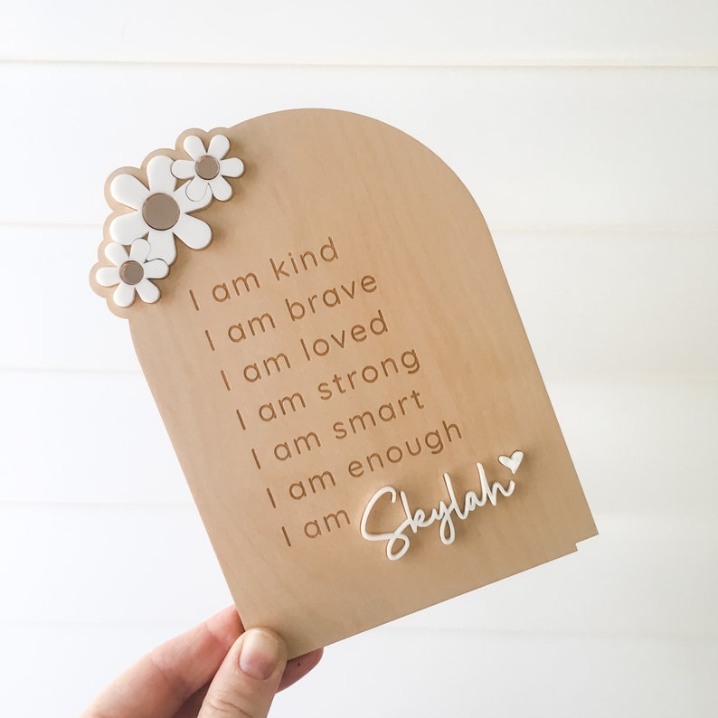 Puede incluir: Un letrero de madera con un fondo marr&oacute;n claro y flores blancas. El letrero tiene el texto "Soy amable, soy valiente, soy amada, soy fuerte, soy inteligente, soy suficiente, soy Skylah" grabado en &eacute;l.