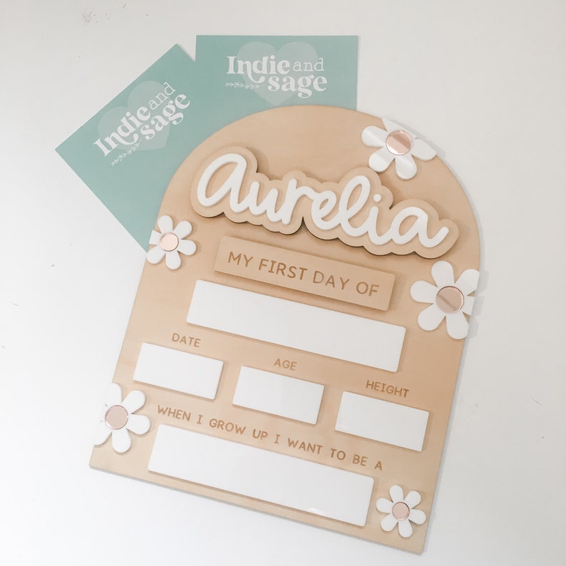 Op de afbeelding: Een houten bord met de naam "Aurelia" in witte letters, versierd met witte madeliefjes en de tekst "MY FIRST DAY OF." Het heeft vakken voor datum, leeftijd, lengte en ambities. Twee lichtblauwe kaarten met "Indie and Sage" zijn op de achtergrond.