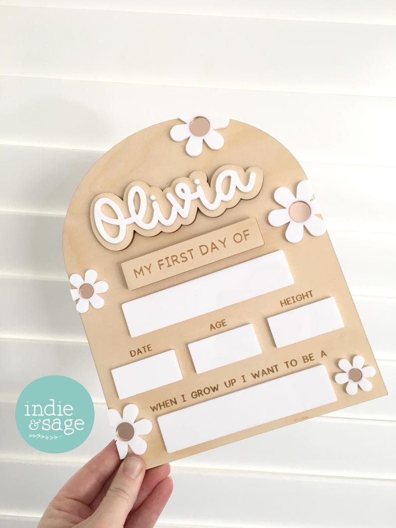 Op de afbeelding: Houten bord voor de eerste schooldag met de naam "Olivia" in witte letters. Het bord heeft een lichtbruine achtergrond, witte madeliefjesaccenten en invulvakken.